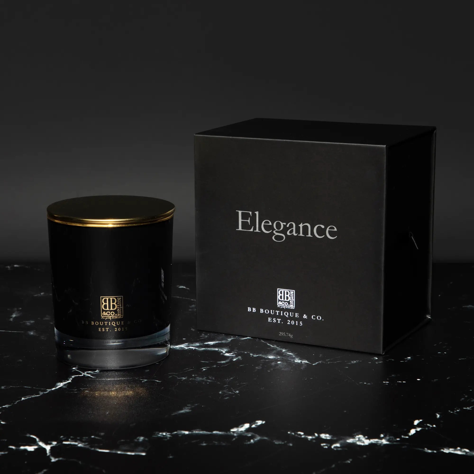 Photograph of a BB Boutique & Co. Elegance luxury candle in a black glass jar with a gold lid and its matching black box featuring elegant silver text and gold branding, resting on a striking black marble surface with white veining