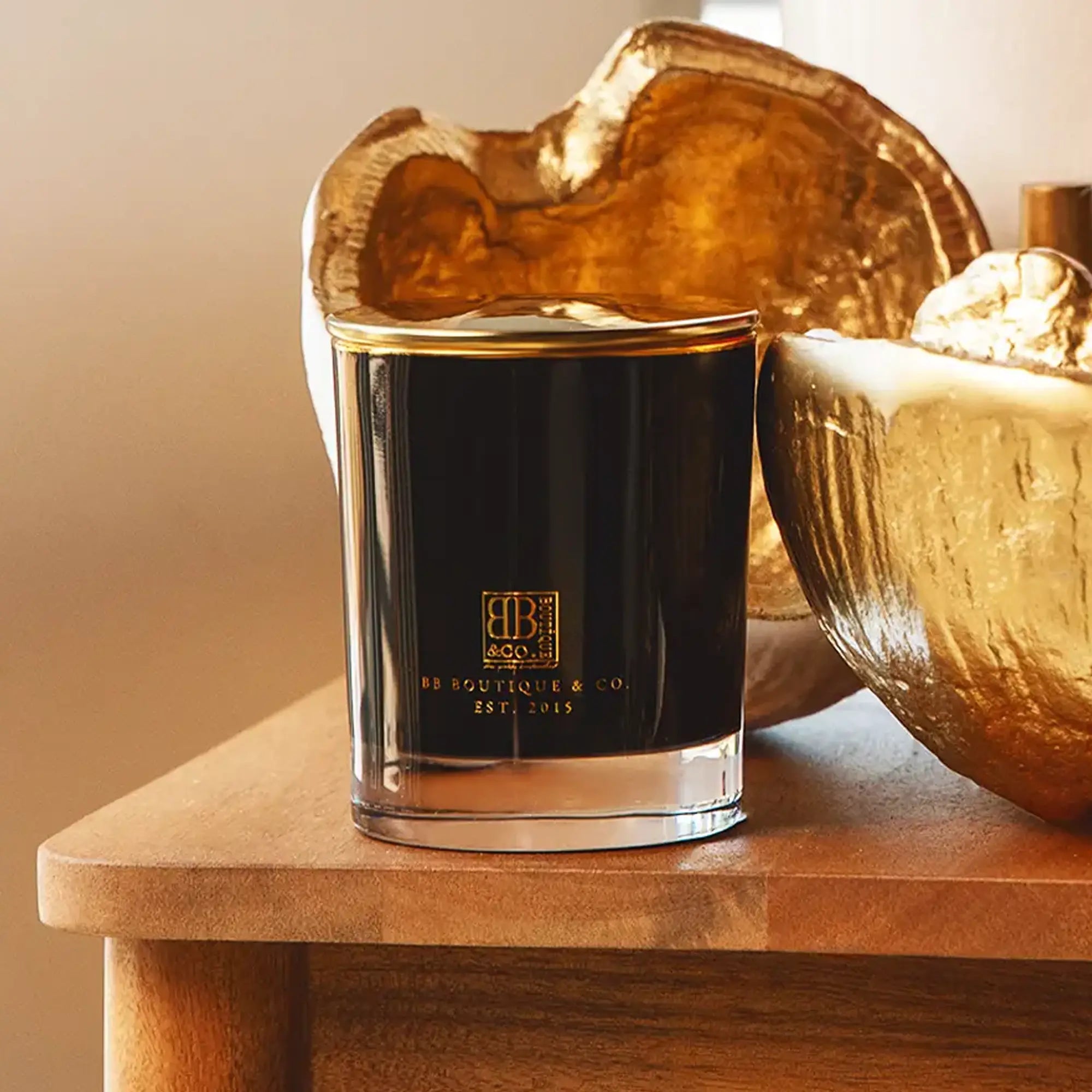 A black BB Boutique & Co. candle with gold lettering and trim on a rustic wooden table, flanked by textured golden decorative bowls