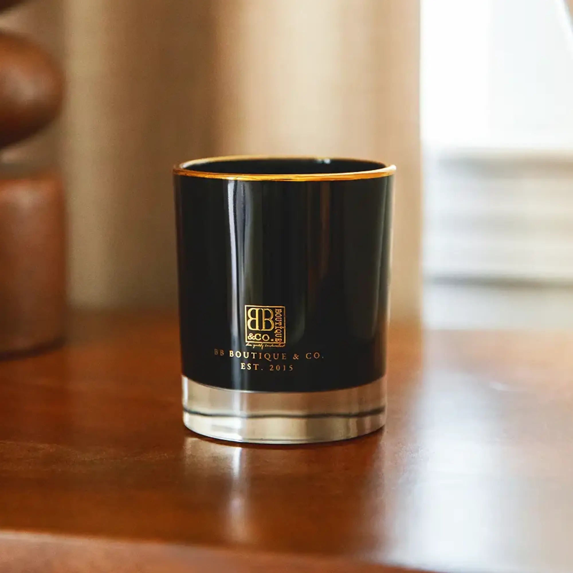 Photograph of a BB Boutique & Co. candle in a glossy black jar with gold trim and logo and a reflective silver base, displayed on a polished wooden table