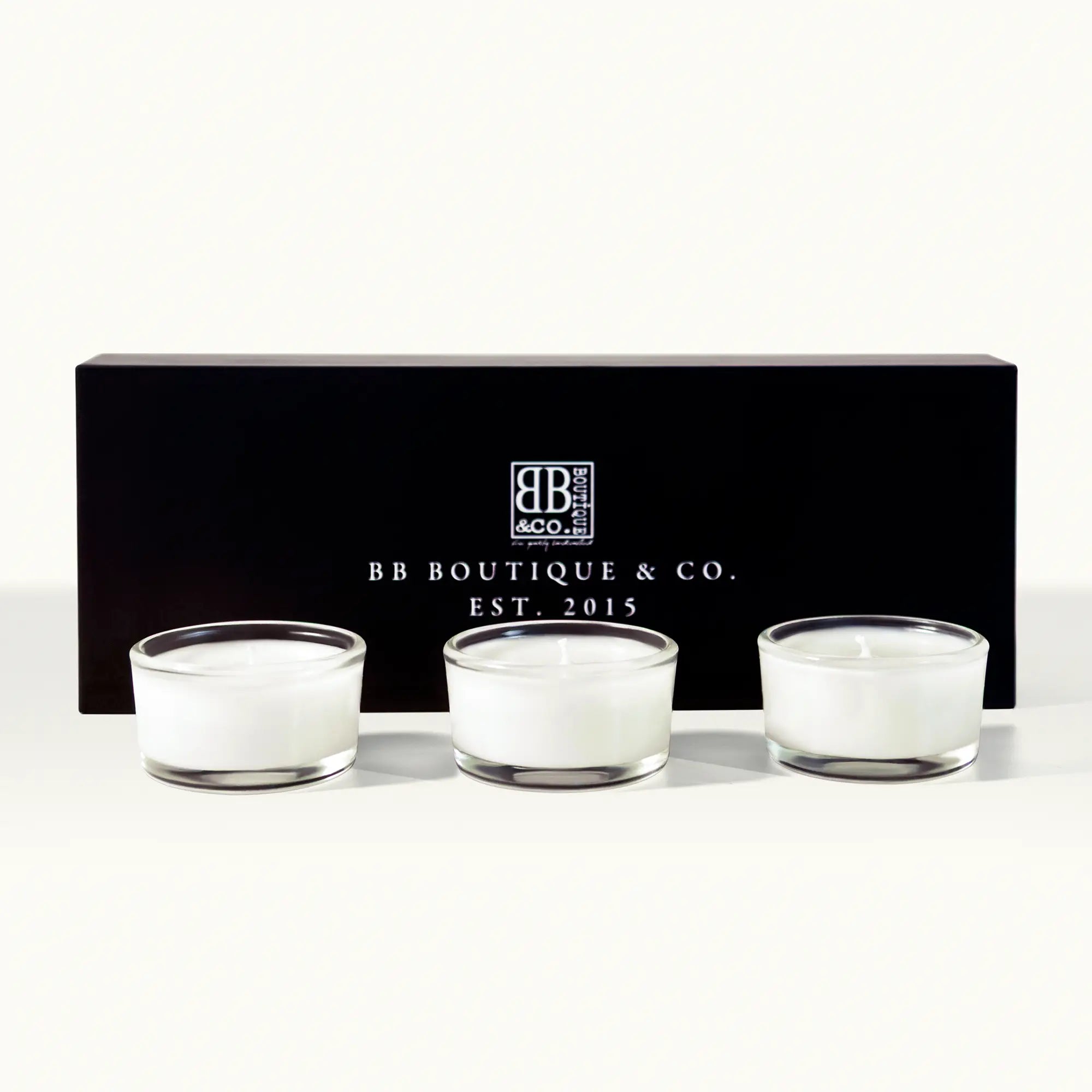Product photography of three white BB Boutique & Co. votive candles in clear glass holders positioned in front of a sleek black gift box displaying the "BB Boutique & Co. EST. 2015" logo in white on a clean white studio background