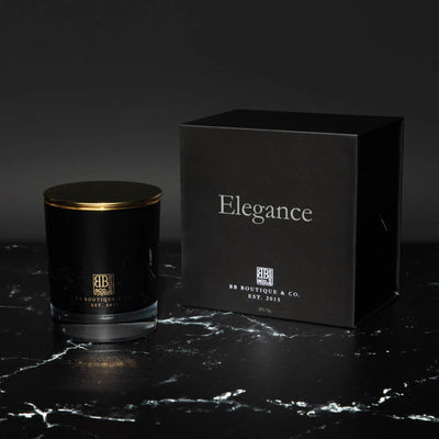 Photograph of a BB Boutique & Co. Elegance luxury candle in a black glass jar with a gold lid and its matching black box featuring elegant silver text and gold branding, resting on a striking black marble surface with white veining