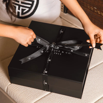 A person's hands untying a black satin ribbon with "BB Boutique & Co. EST. 2015" branding from an elegant black gift box on a cream textured surface