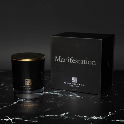 Commercial photograph featuring a black candle with a gleaming gold lid and a matching black box labeled "Manifestation" and "BB BOUTIQUE & CO. EST. 2015", all displayed on a luxurious black marble surface with intricate white veining