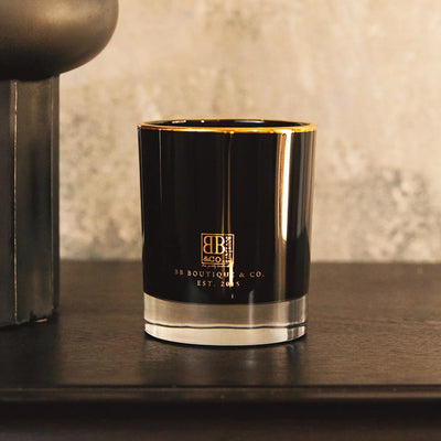 BB Boutique & Co. luxury candle featuring a sleek black glass vessel with a gold rim, clear base, and a prominent gold BB & Co. logo, placed on a dark wooden surface