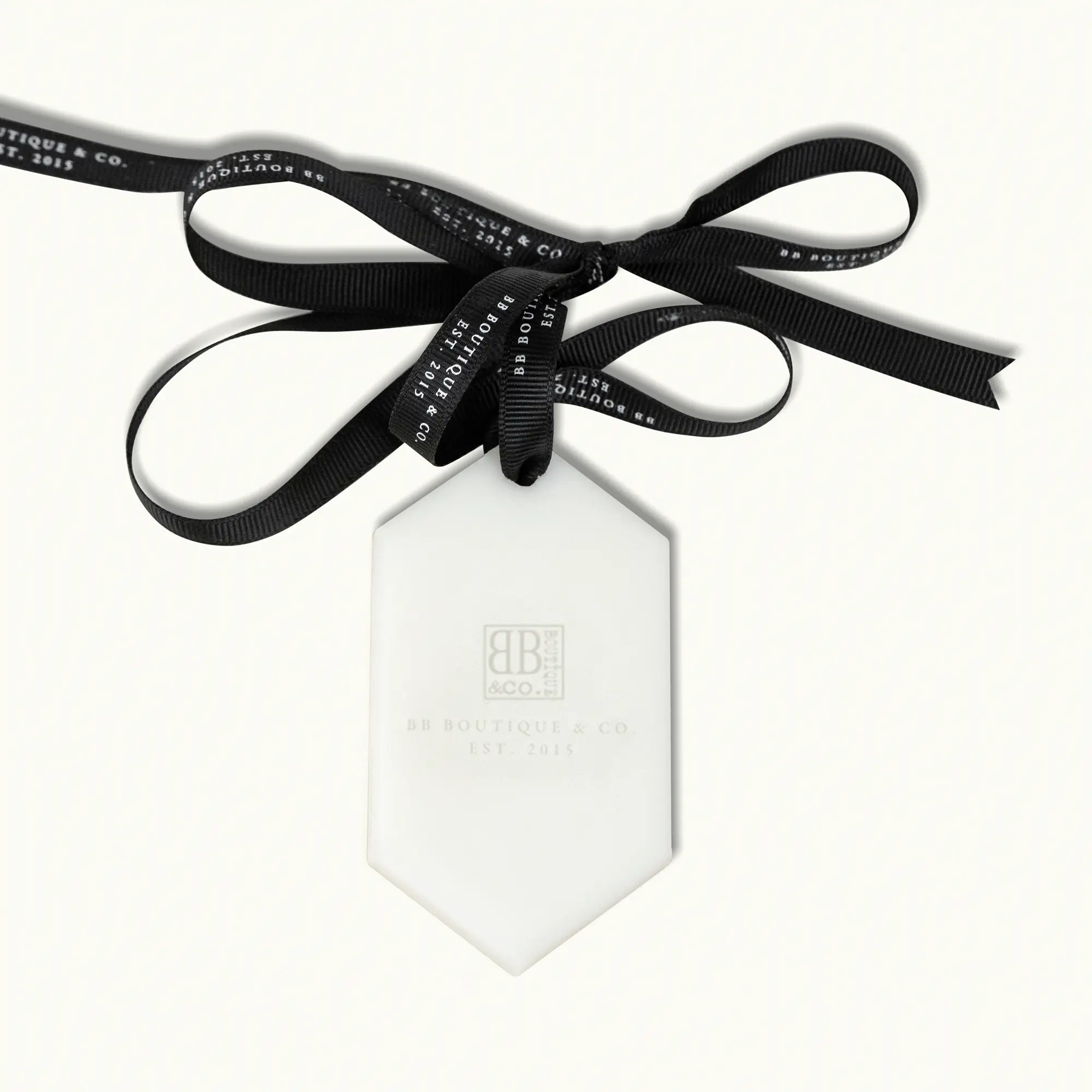 White hexagonal BB Boutique & Co brand tag with a subtle grey logo and text hanging from a black grosgrain ribbon tied in a bow with repeating white BB Boutique & Co Est 2015 text against a clean white background