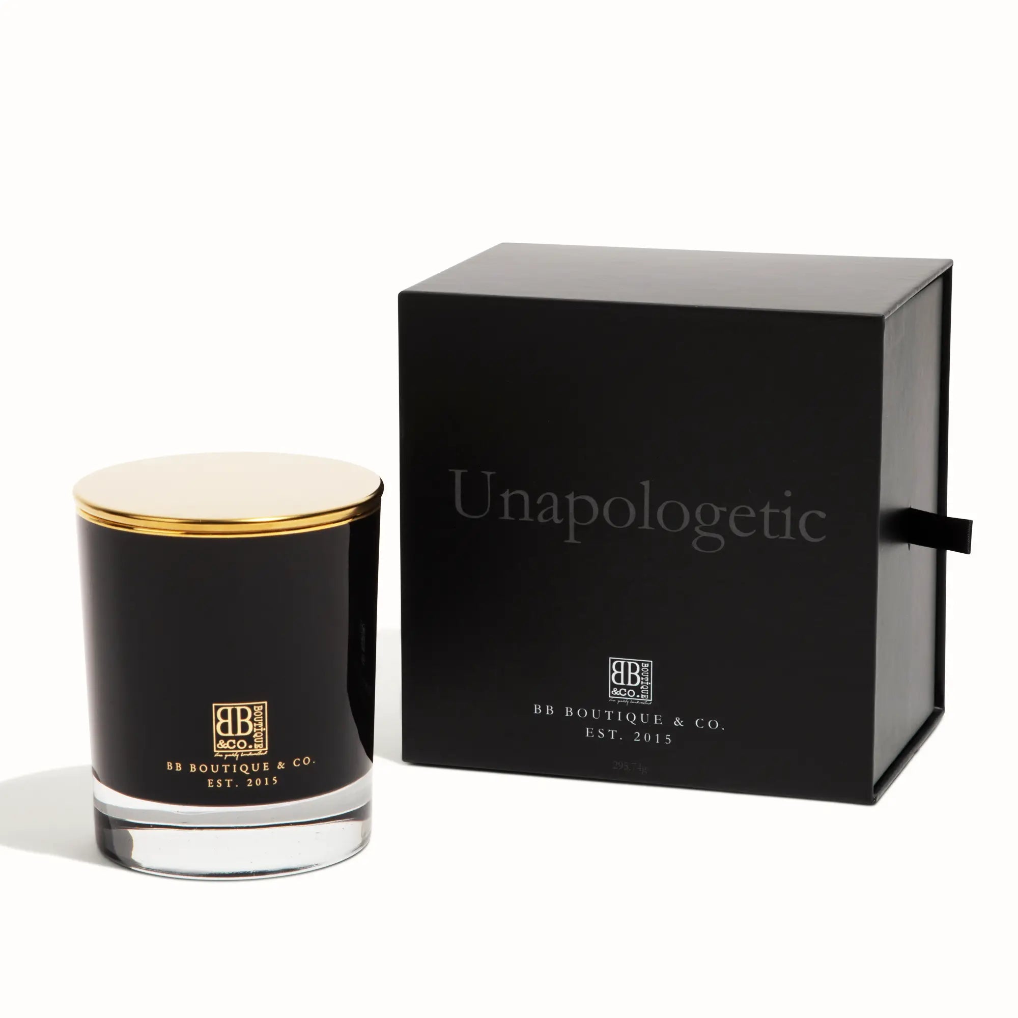 BB Boutique & Co. product photography of an Unapologetic luxury candle, featuring a sleek black glass vessel with a gold lid and matching branding, displayed next to its elegant black gift box with bold "Unapologetic" text and a black ribbon pull tab