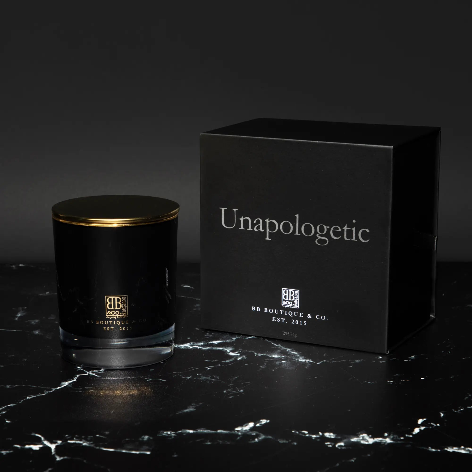 Luxury product shot of a black glass candle jar with a gold lid and a matching black box featuring silver "Unapologetic" text and the BB Boutique & Co. logo on a black marble surface with elegant white veining