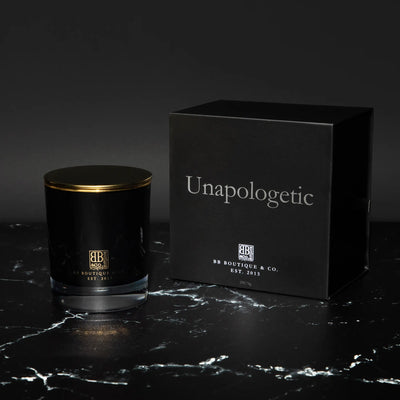 Luxury product shot of a black glass candle jar with a gold lid and a matching black box featuring silver "Unapologetic" text and the BB Boutique & Co. logo on a black marble surface with elegant white veining