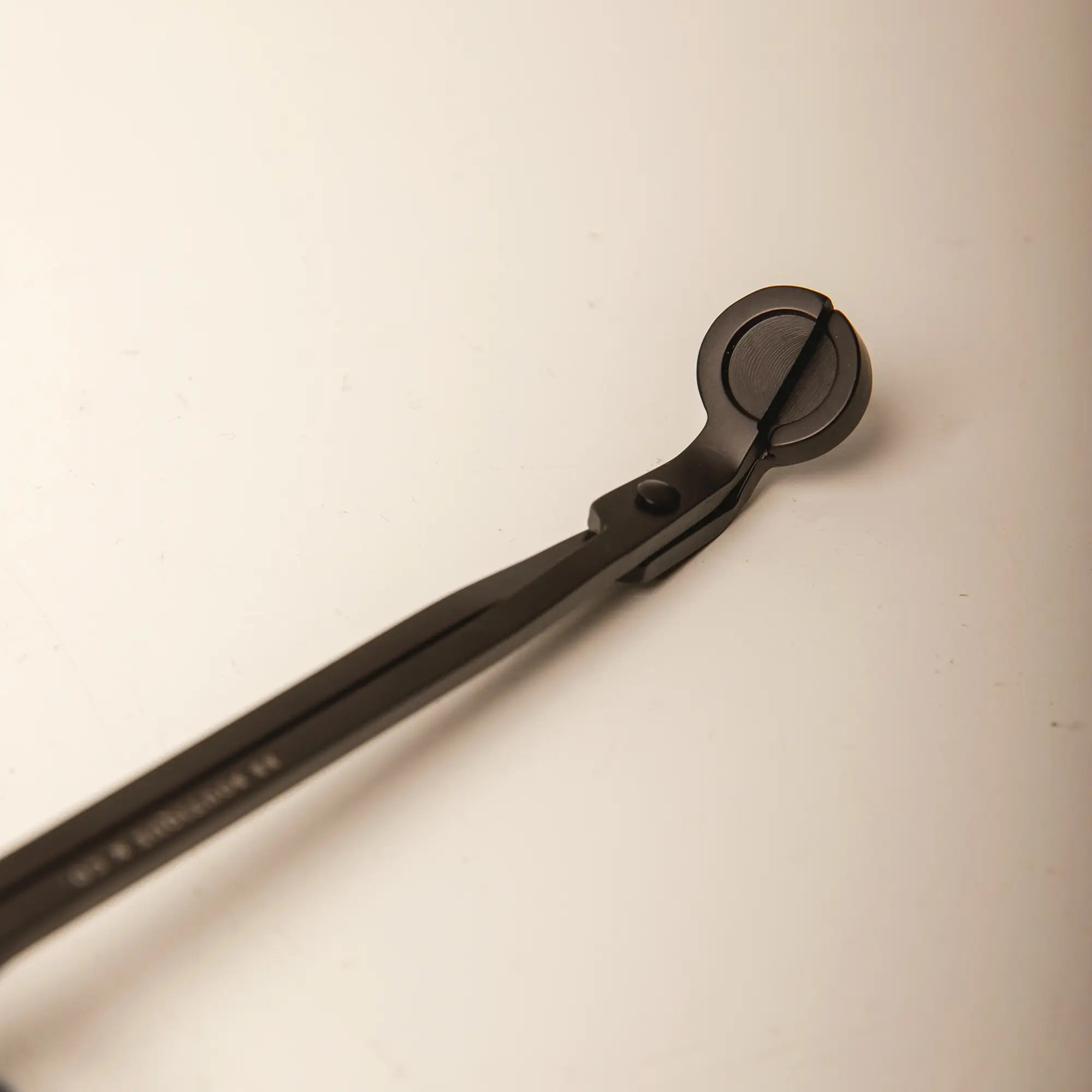A close-up photograph of a matte black candle wick trimmer with a circular cutting head and slender handles angled across a plain light beige background