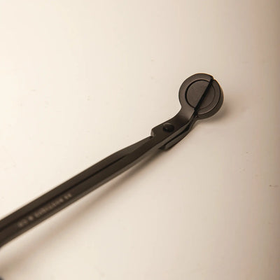 A close-up photograph of a matte black candle wick trimmer with a circular cutting head and slender handles angled across a plain light beige background
