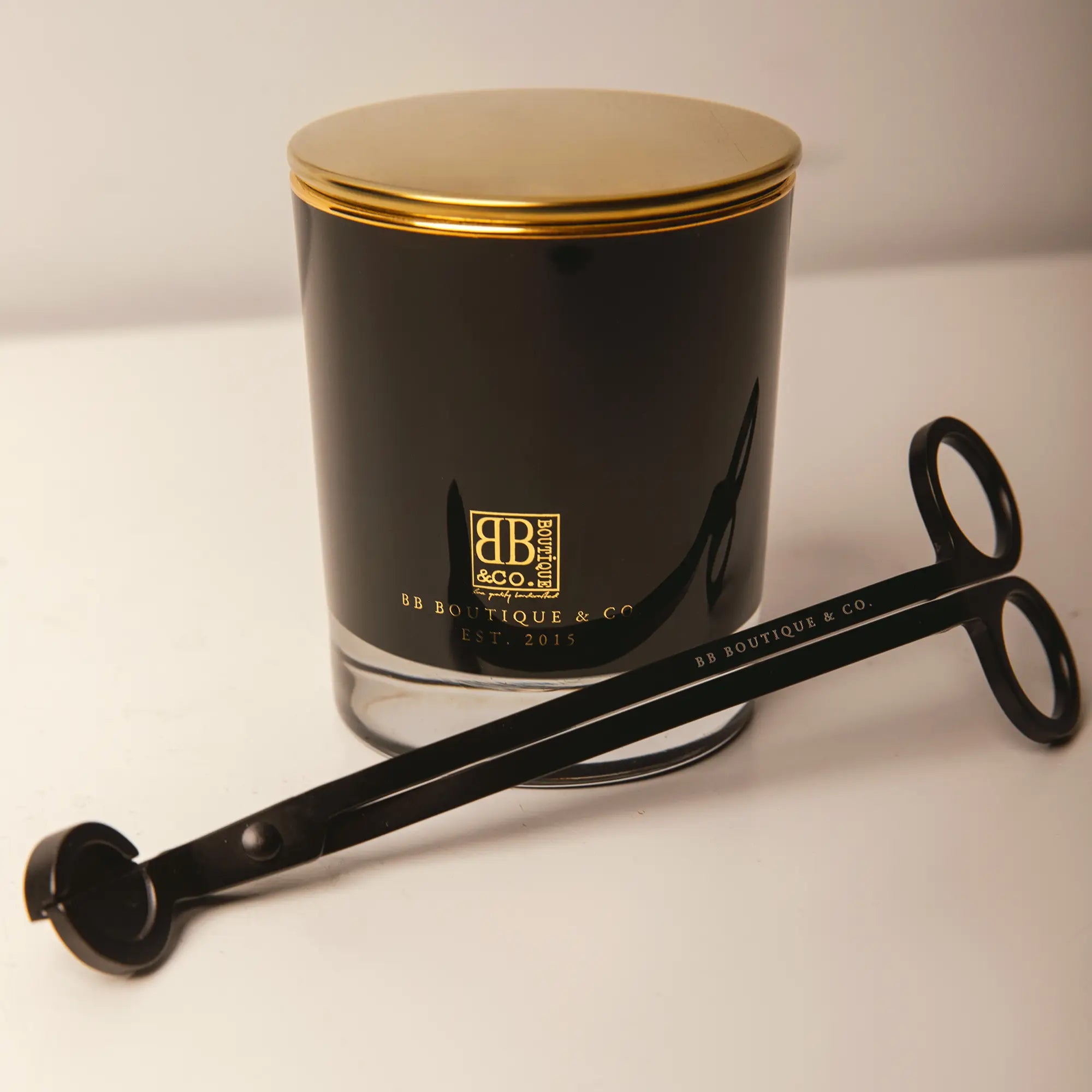 BB Boutique & Co. product photography showcasing a luxurious black candle jar with a shining gold lid and prominent gold brand logo next to a matching matte black wick trimmer on a clean white studio background