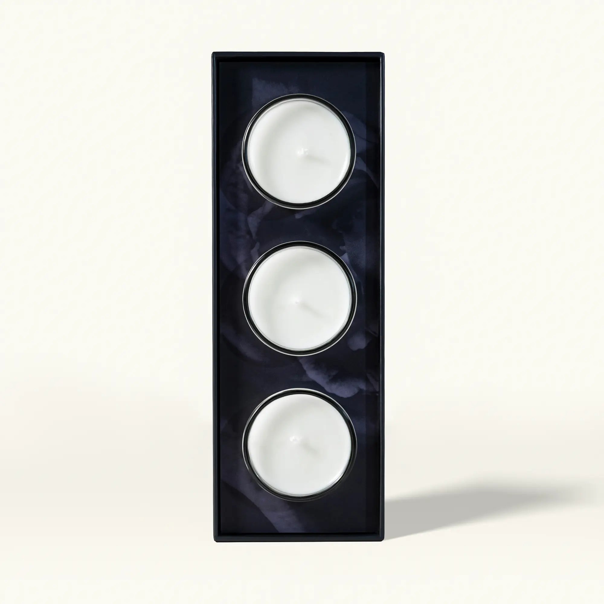 A set of three white tea-light candles arranged vertically in a black rectangular holder featuring a subtle dark floral pattern on a plain light background