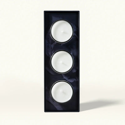 A set of three white tea-light candles arranged vertically in a black rectangular holder featuring a subtle dark floral pattern on a plain light background