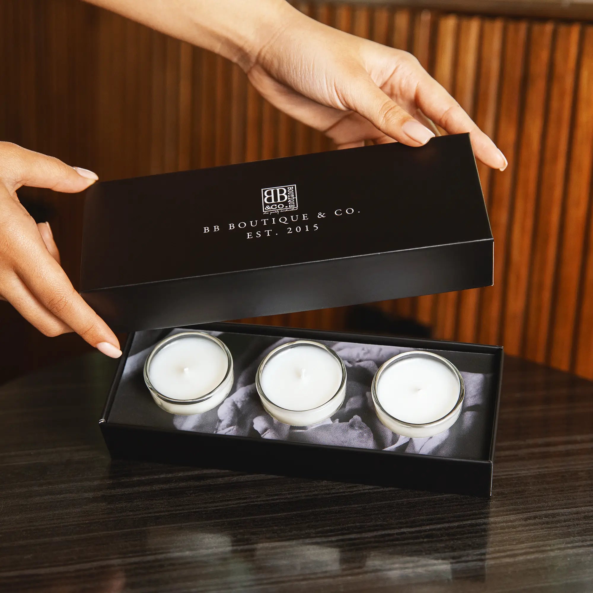 Photography of a person's hands opening a sleek black BB Boutique & Co. gift box revealing three white wax candles nestled in grey filler on a dark wood table
